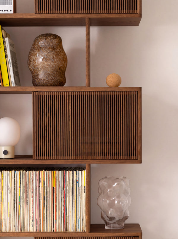 MOLL in solid walnut, close-up of a slatted front between shelves holding records, a glass vase and a table lamp