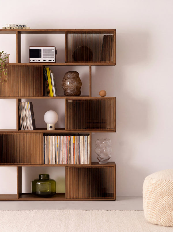 Angled view of MOLL in solid walnut, with staggered shelves, slatted fronts, decor and a pouf alongside