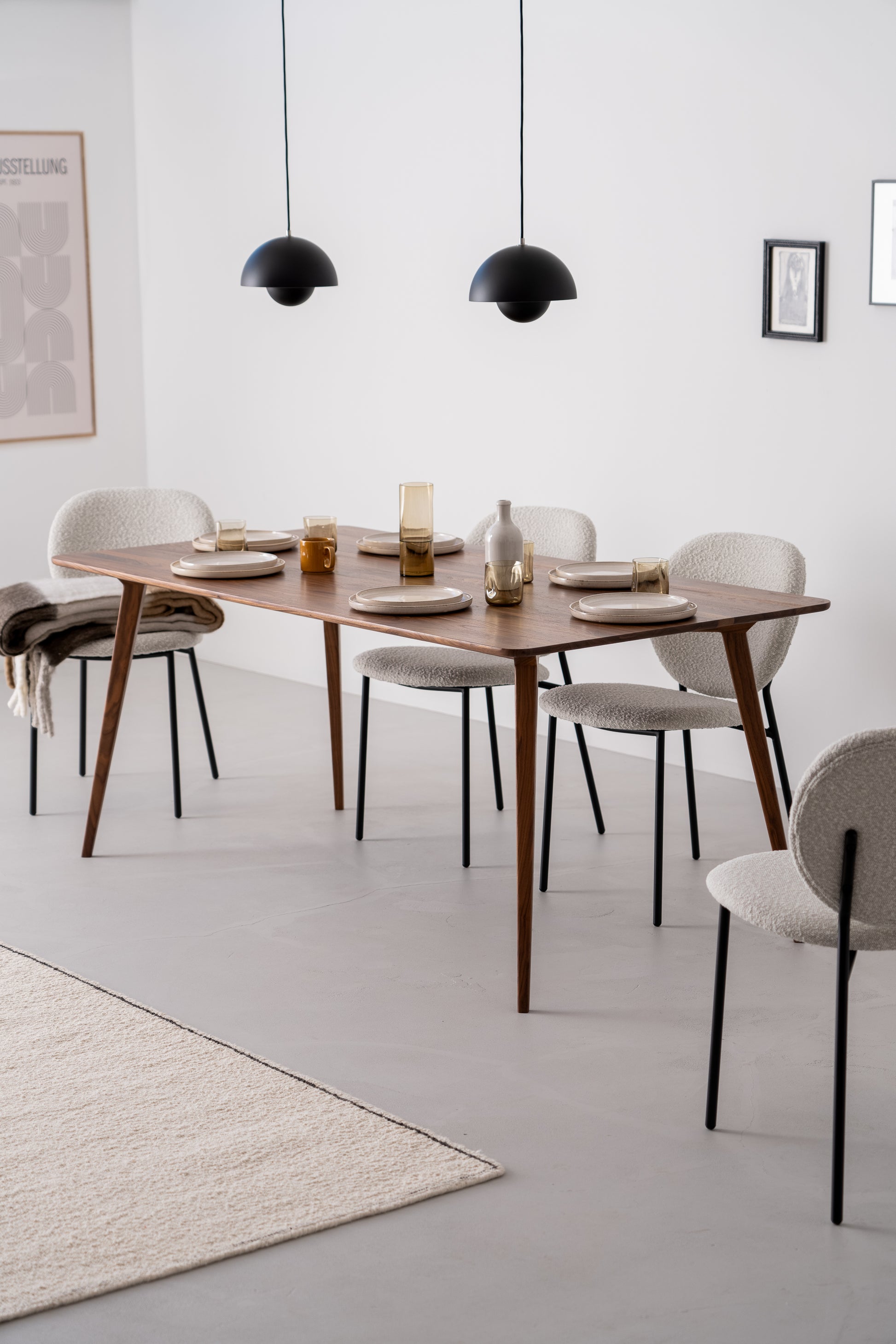 A minimalist dining room with a wooden table, four white chairs, black pendant lights, and simple table settings.