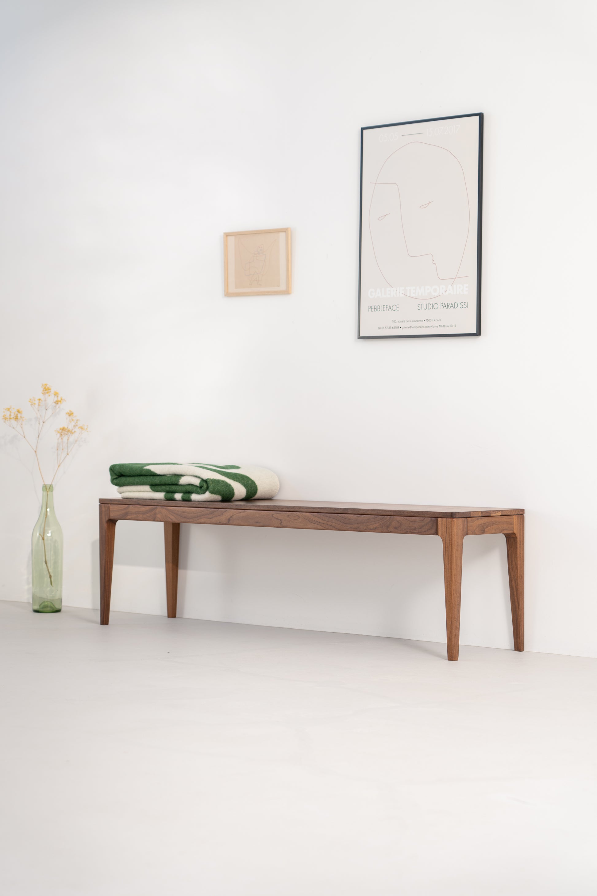 Minimalist room with a wooden bench, folded blanket, wall art, and a tall vase with dried flowers.
