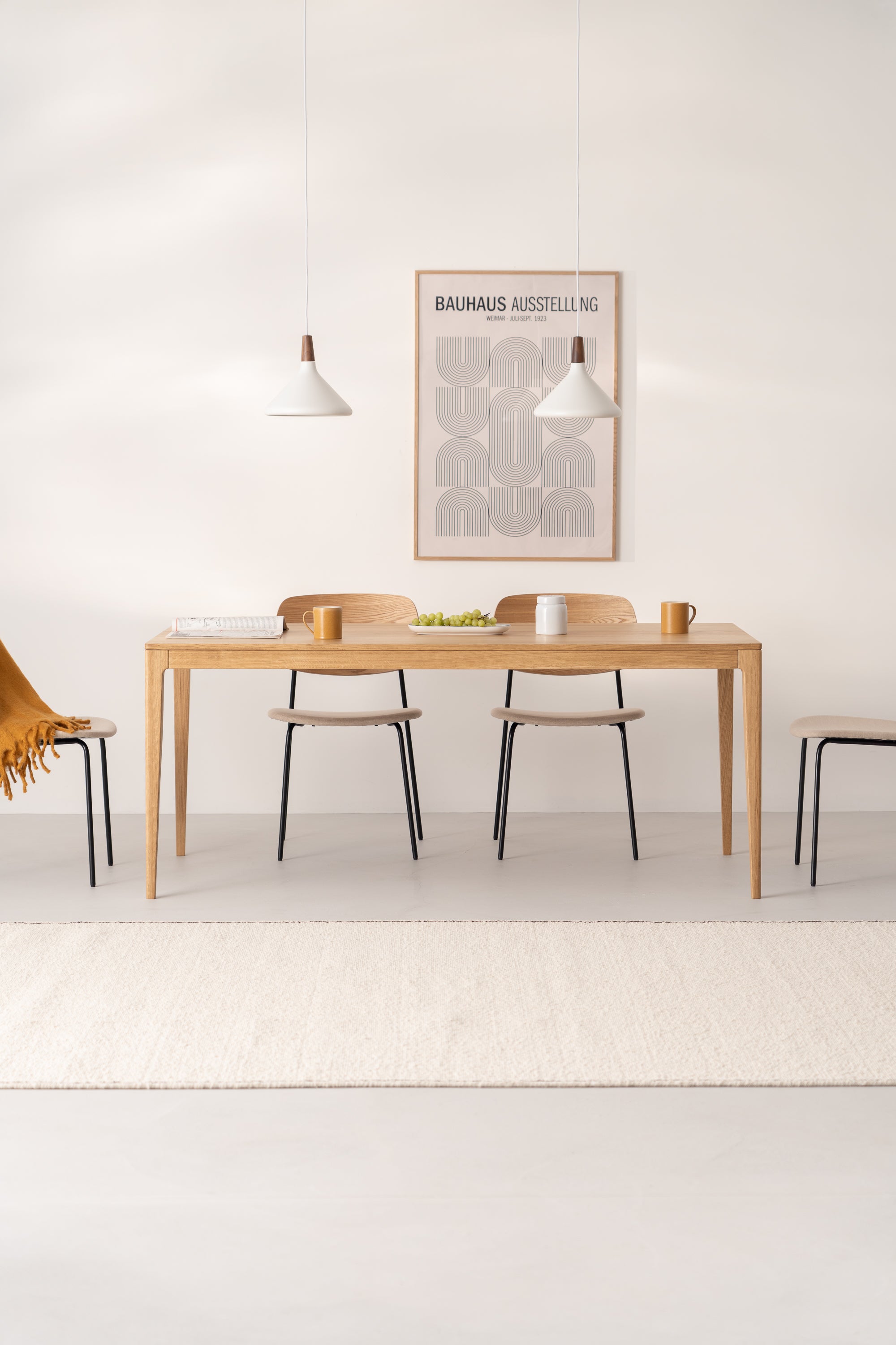 Minimalist dining room with wooden table, three chairs, wall art, pendant lights, and a white rug.