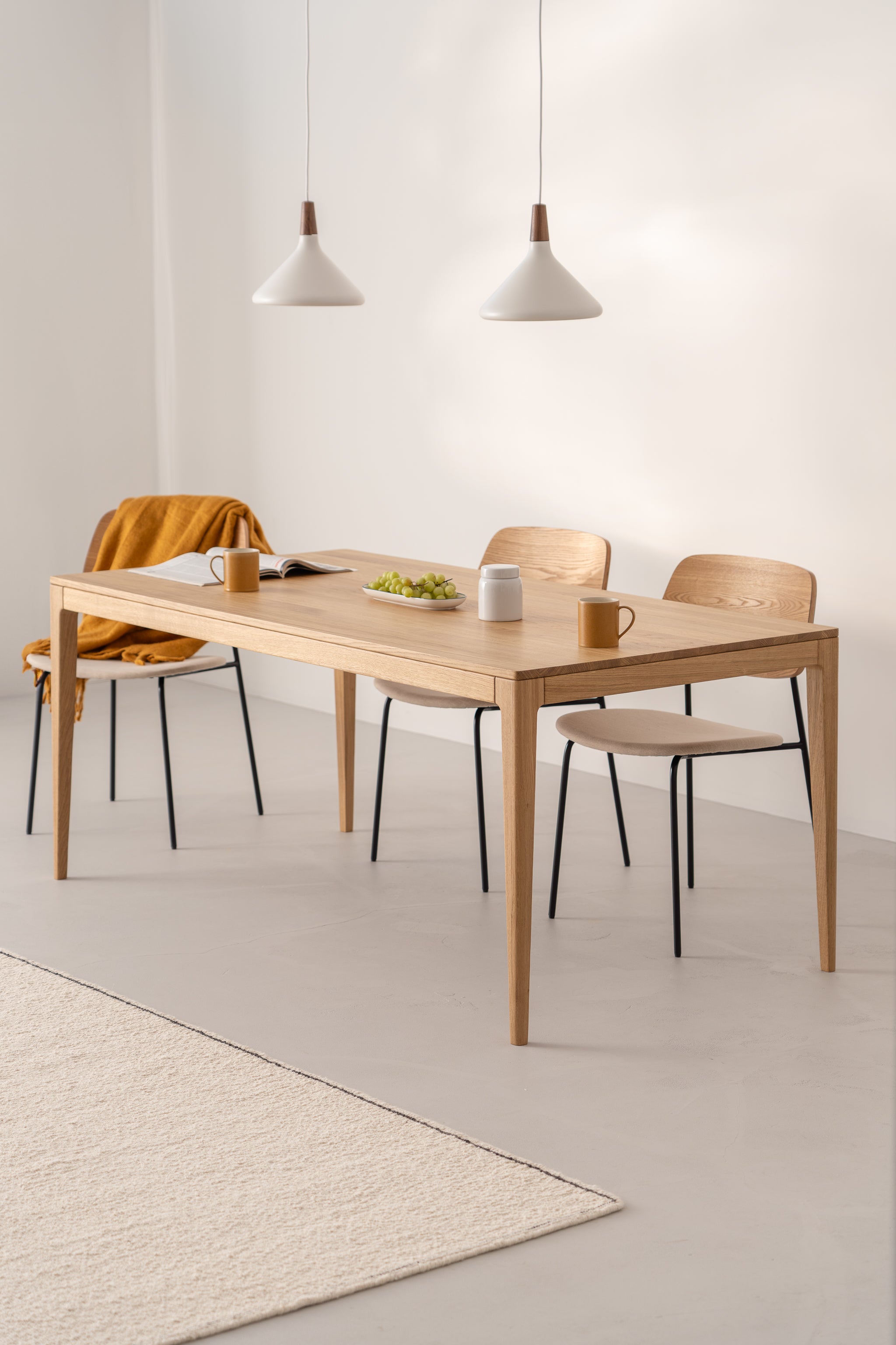 Minimalist dining area with a wooden table, two chairs, pendant lights, a mustard throw, and a plate of grapes.