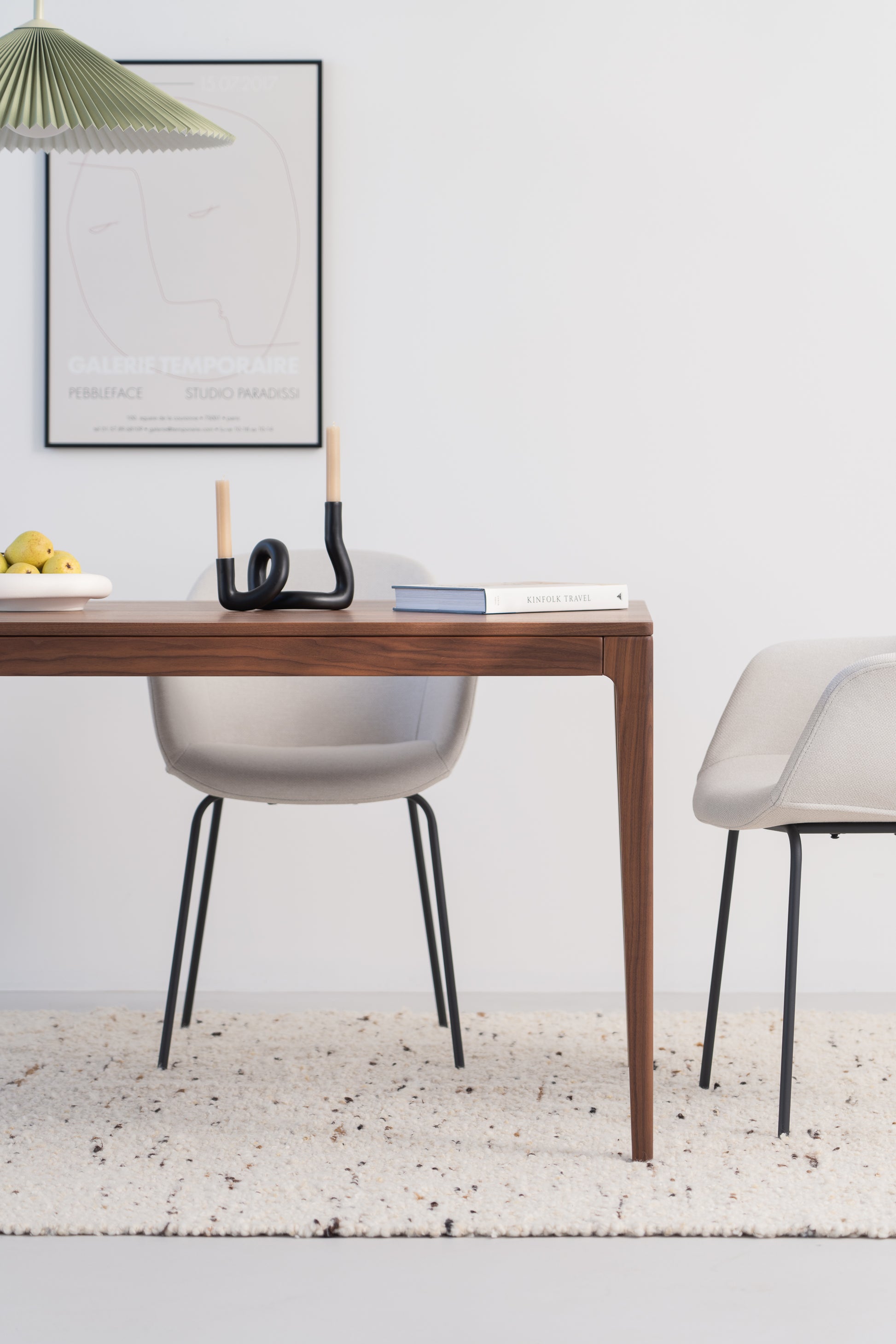 Minimalist dining area with a wooden table, two chairs, fruit bowl, books, and a modern candle holder.