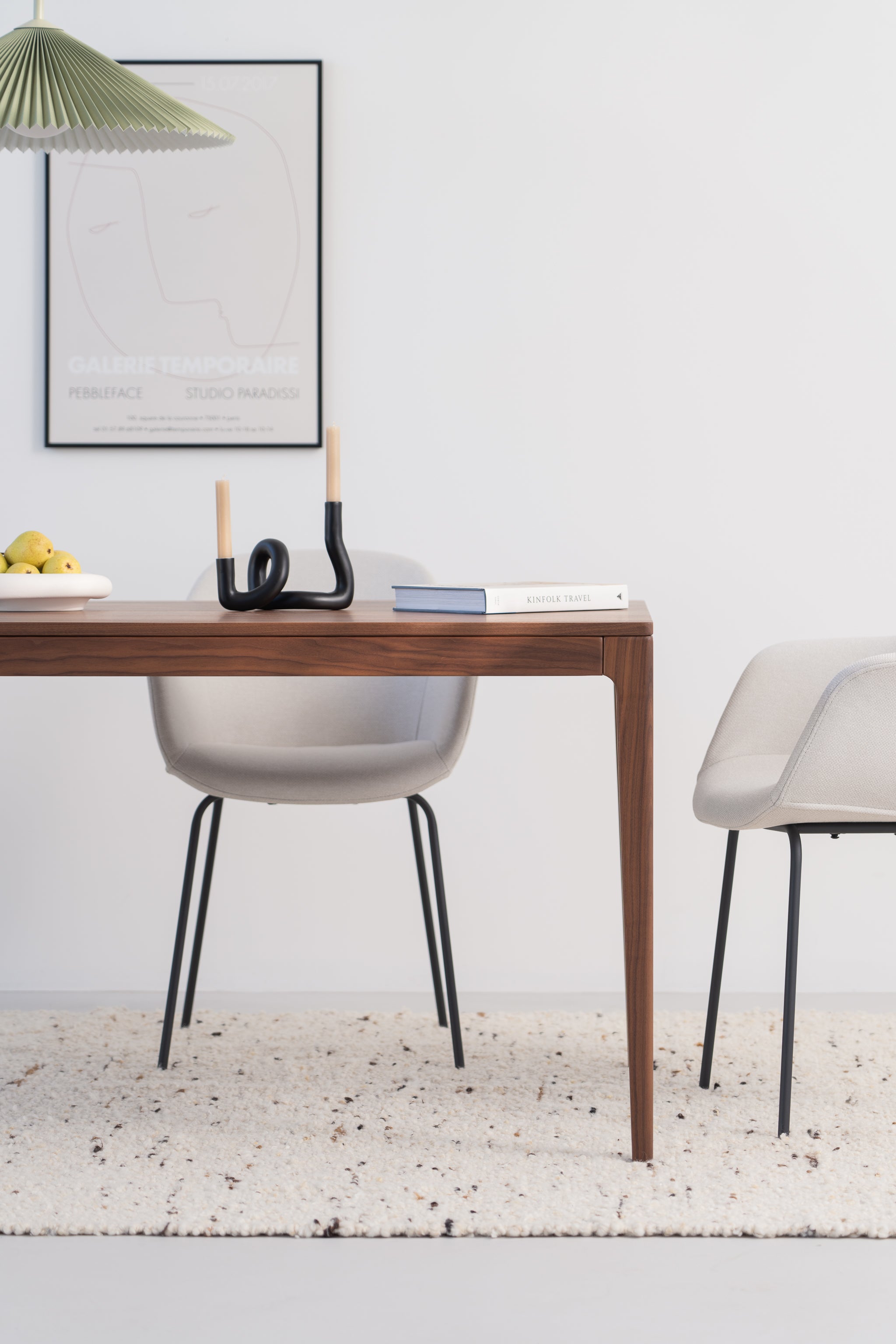 Minimalist dining area with a wooden table, two chairs, fruit bowl, books, and a modern candle holder.