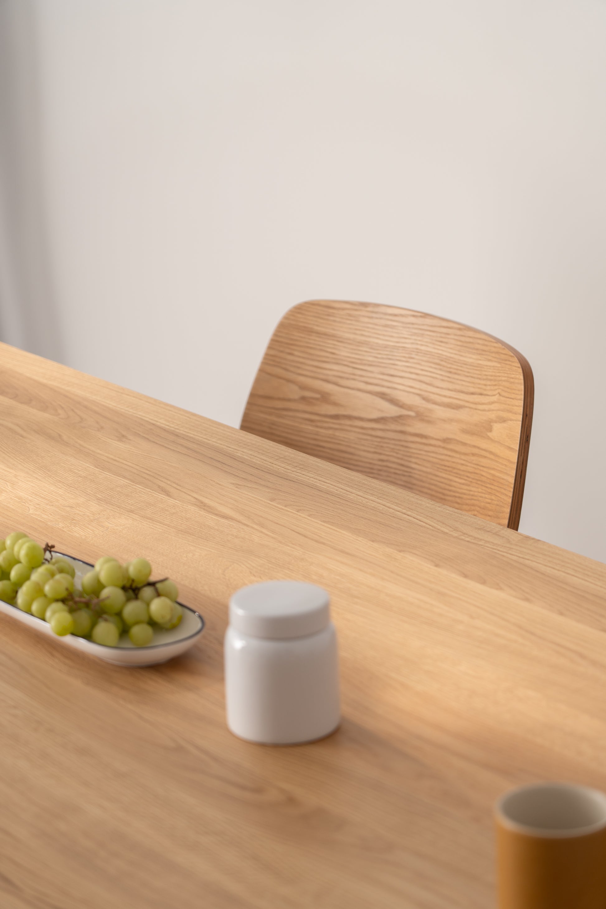 A wooden table with grapes in a dish, a white jar, and a yellow cup, next to a wooden chair.