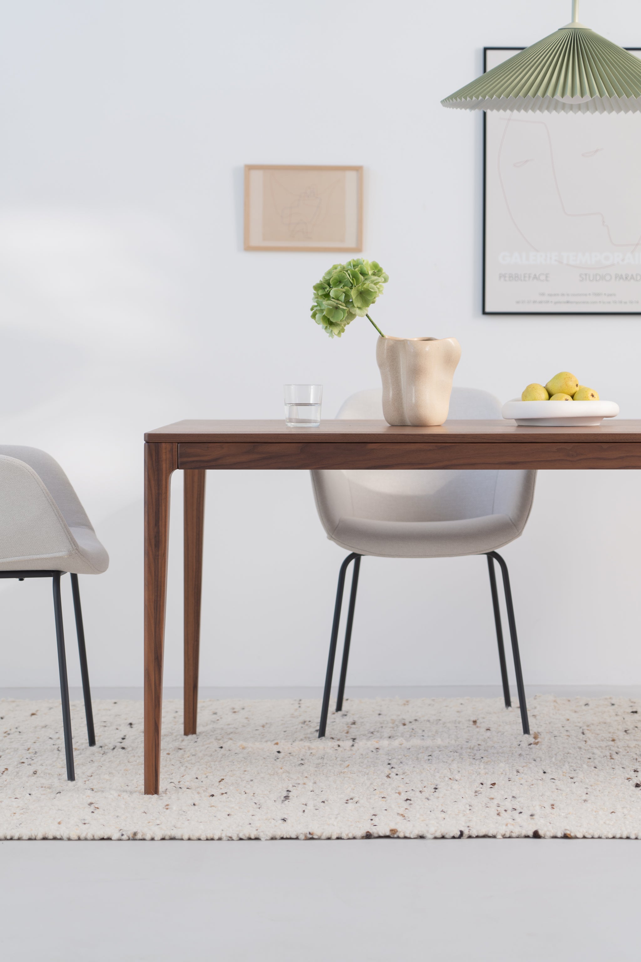 Minimalist dining area with a wooden table, chairs, a vase, and a fruit bowl, set on a textured carpet.