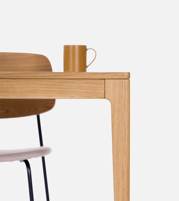 Close-up of a wooden table with a brown mug on top and part of a wooden chair with a cushion seat visible underneath.
