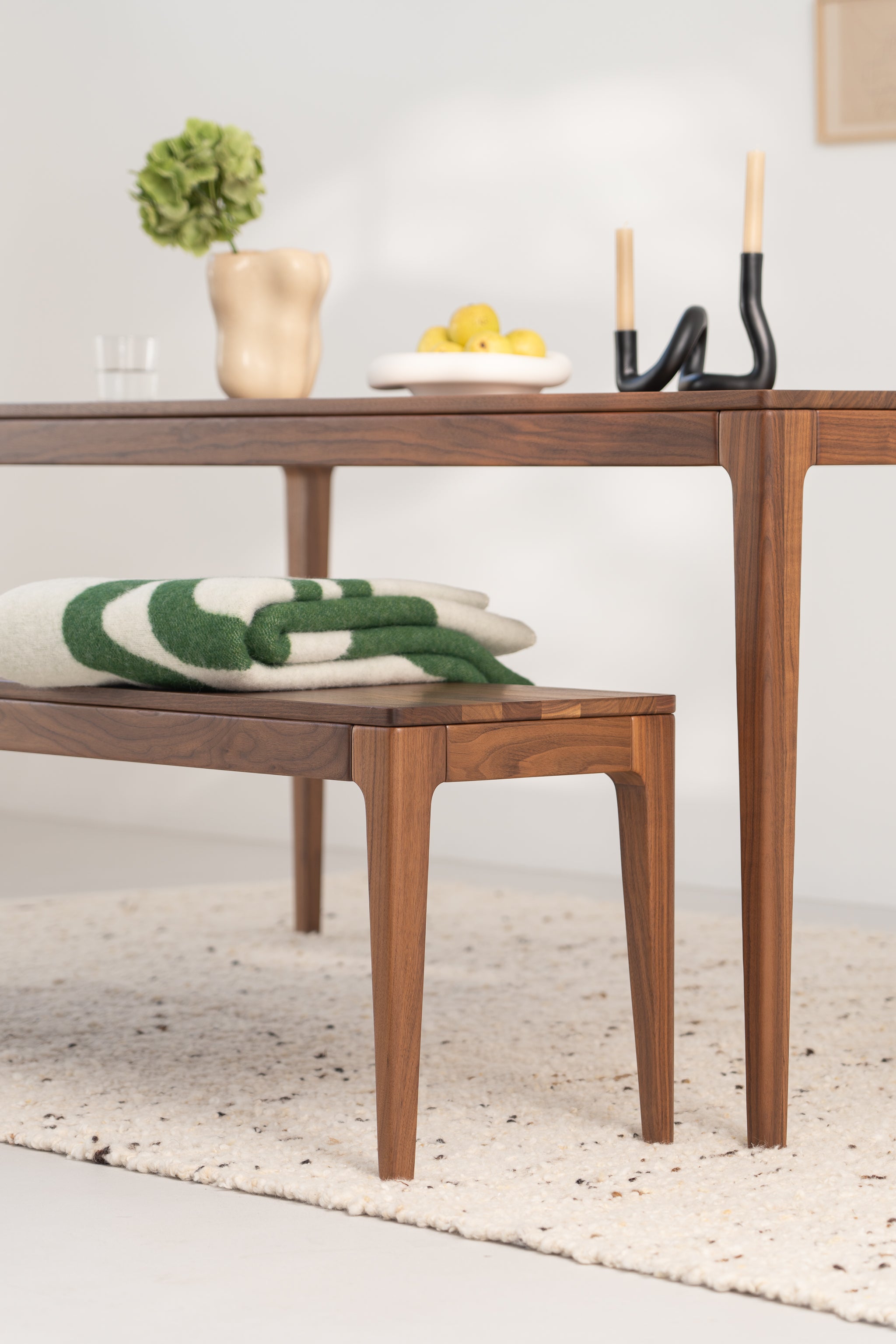 Wooden table and bench with decor items, folded blanket, and fruit bowl in a minimal room setting.
