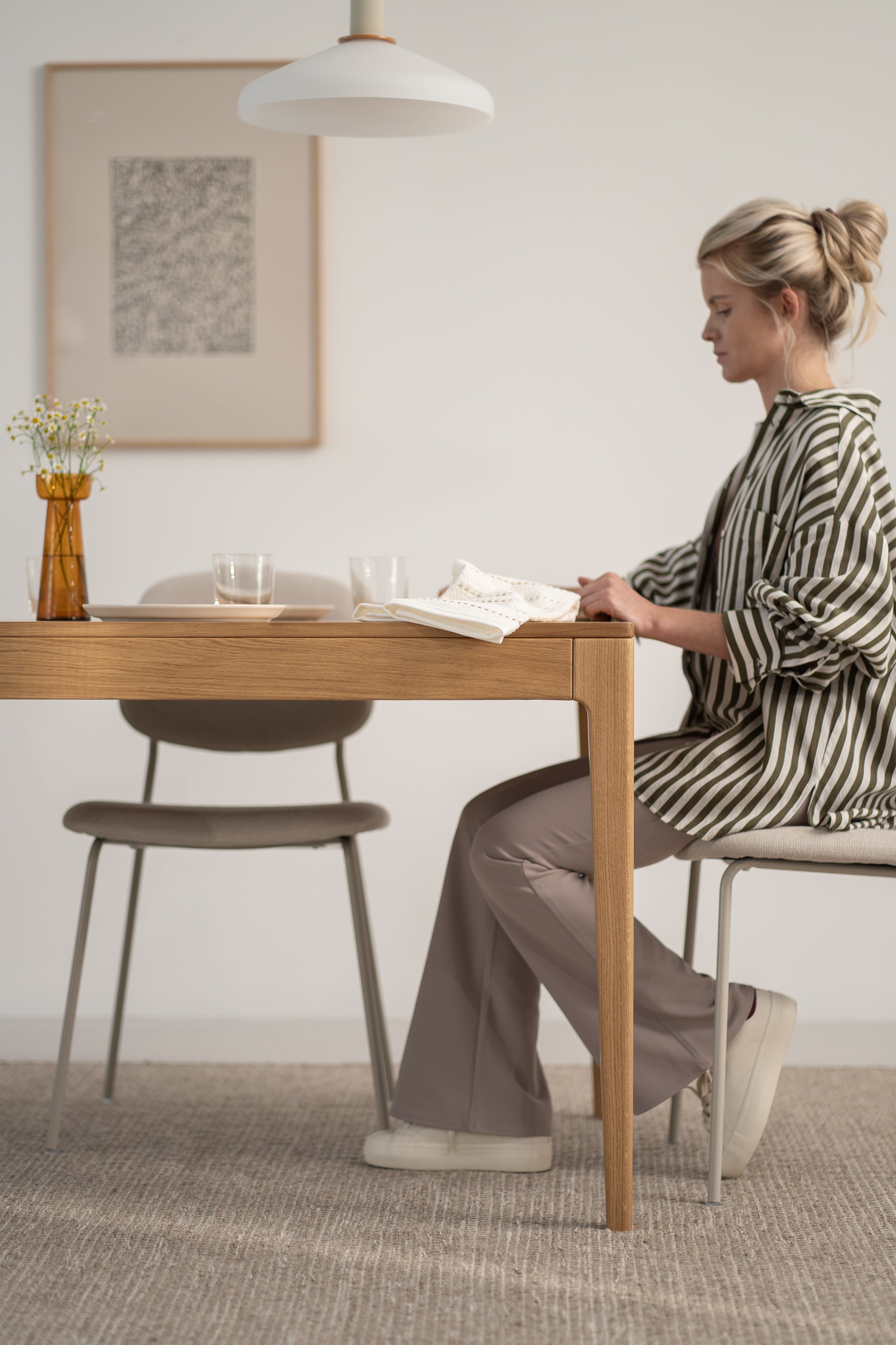 Woman reading at a wooden table in a minimalistic room with a vase and wall art.