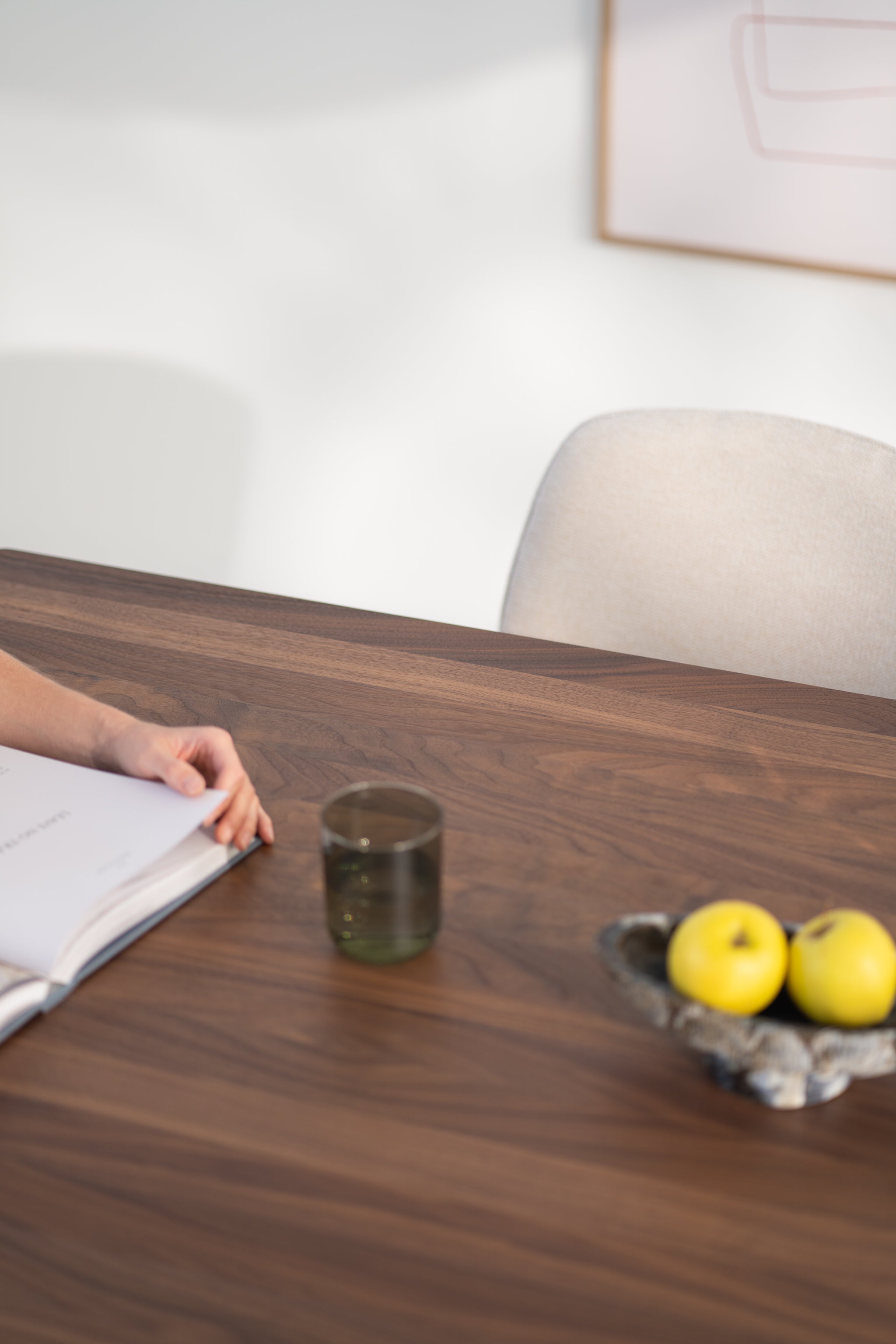 Hand holding a book, glass, and yellow apples on a wooden table.