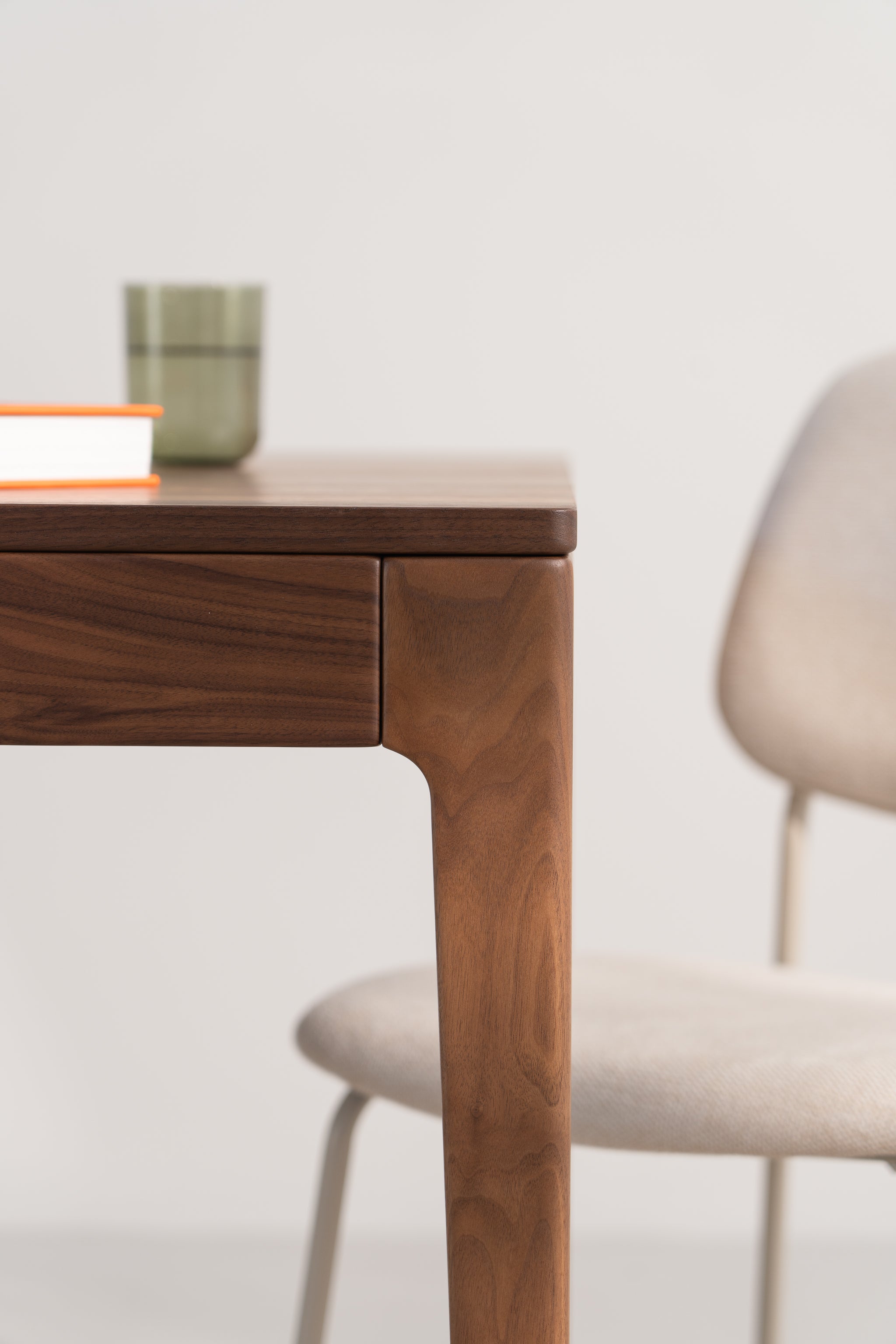 Close-up of a wooden table corner with a red-edged book and a green tumbler, next to a beige chair.