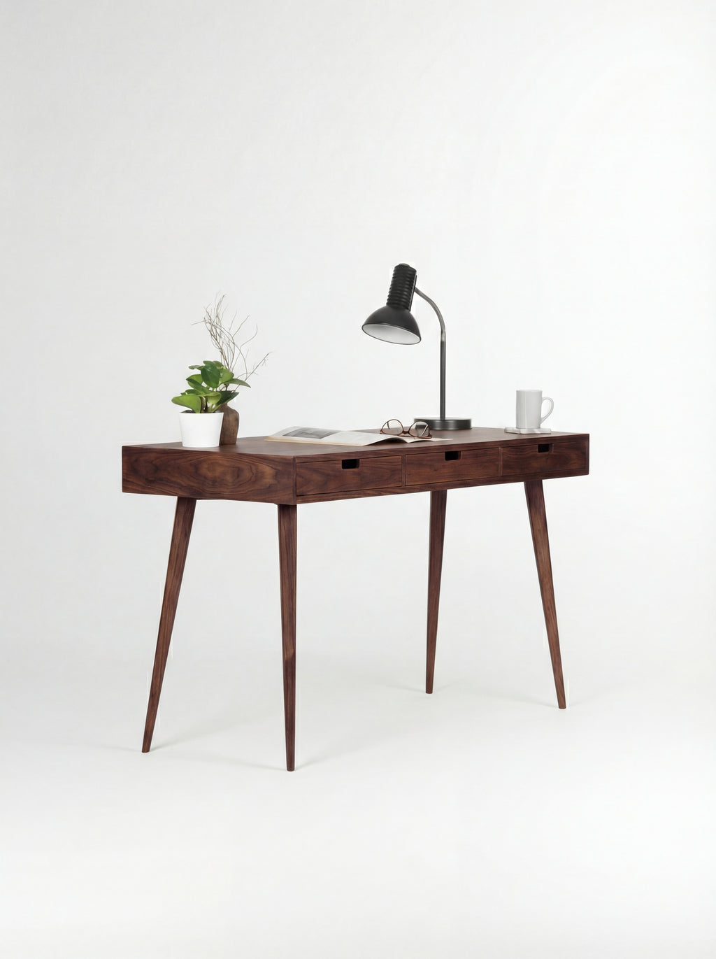 Three-quarter view of the HEIM 140 desk in walnut wood, with a black lamp, plant and open magazine on top