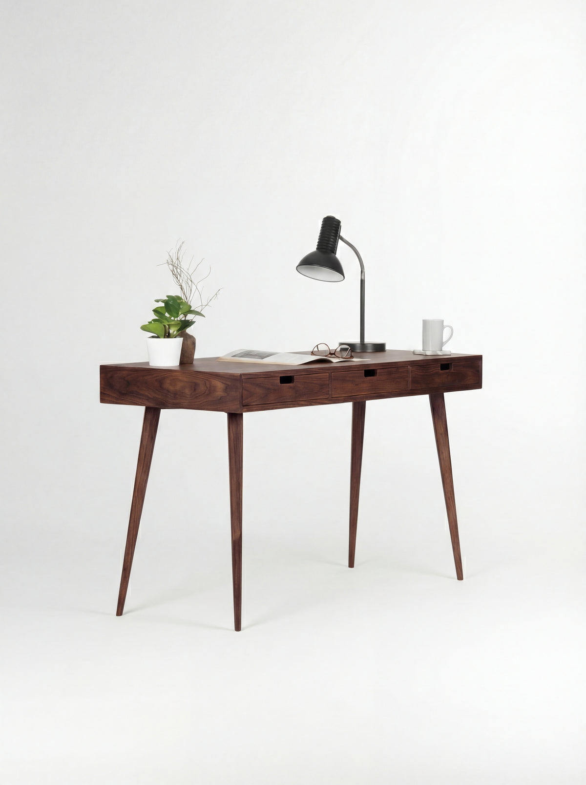 Three-quarter view of the HEIM 140 desk in walnut wood, with a black lamp, plant and open magazine on top