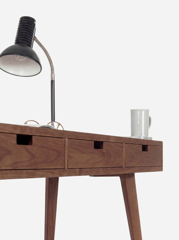 HEIM 140 desk in walnut wood, close-up of three cut-out drawer grips and two legs, with a lamp and mug above