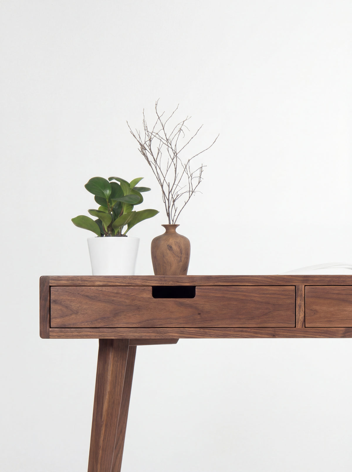 Left corner of the HEIM 140 desk in walnut wood, showing a cut-out drawer grip with a plant and wooden vase above