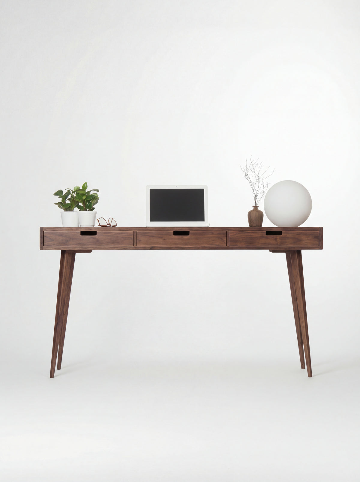 HEIM 140 wooden desk with drawers in walnut wood, shown from the front with a laptop and globe lamp
