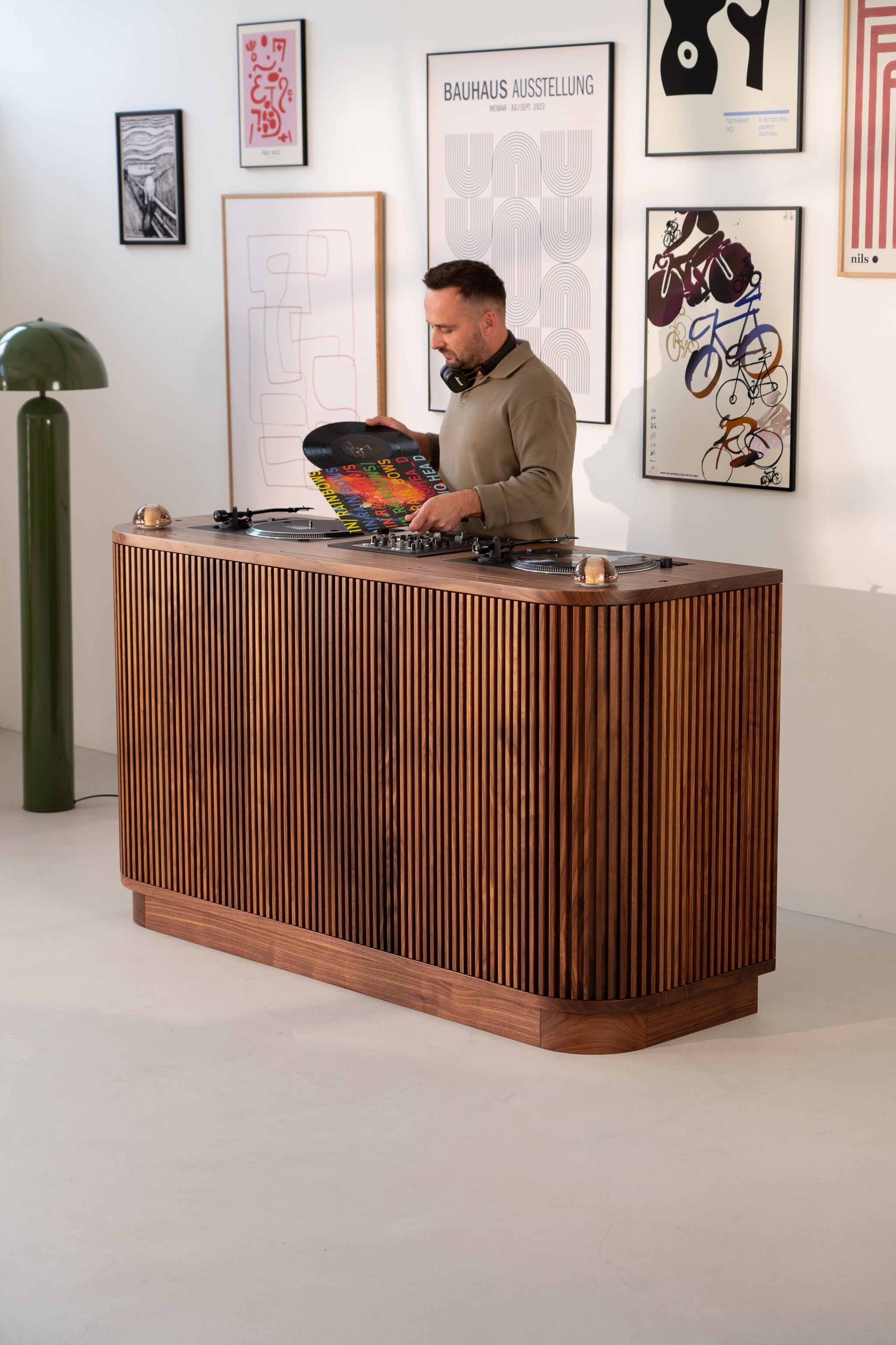 Man standing at a wooden DJ booth holding a vinyl record, with wall art in the background.
