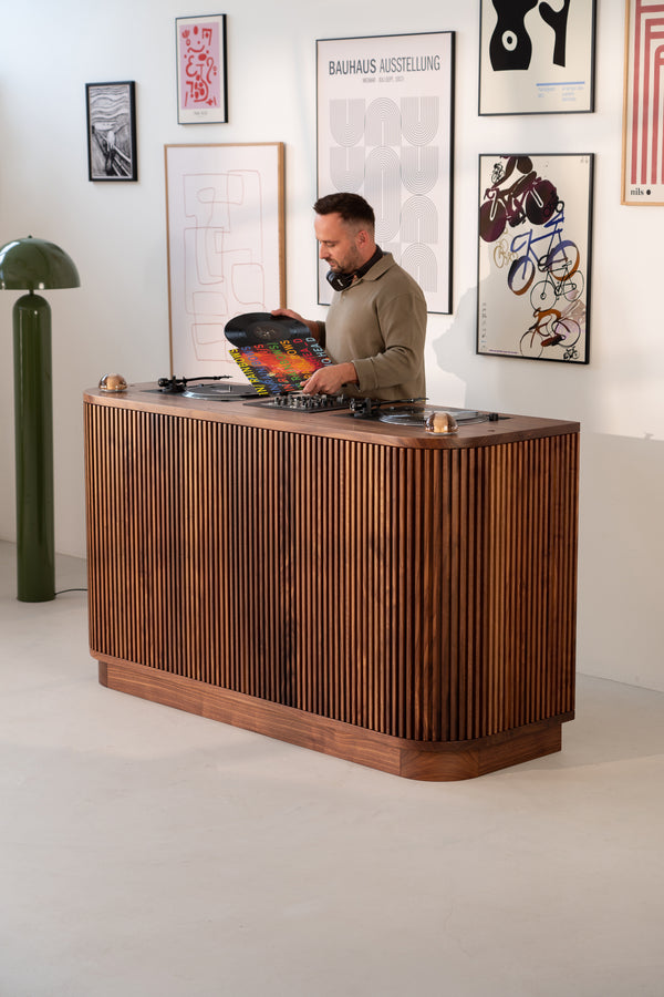 Man standing at a wooden DJ booth holding a vinyl record, with wall art in the background.
