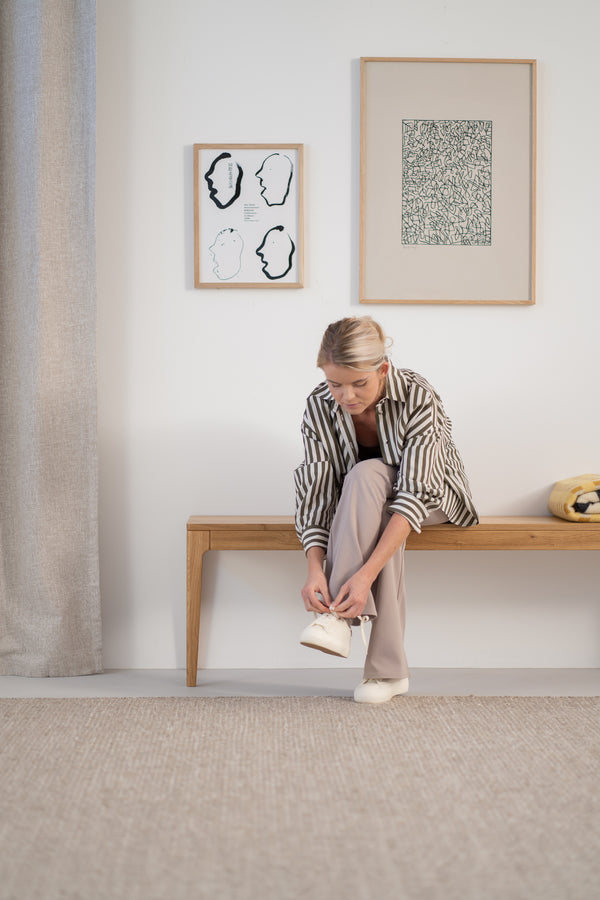 Woman tying shoes on a wooden bench, with art on the wall and a textured rug below.
