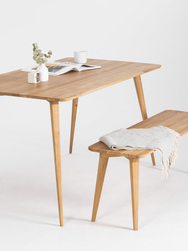 Wooden table and bench on white, with an open magazine, cup, small vase, and folded blanket.
