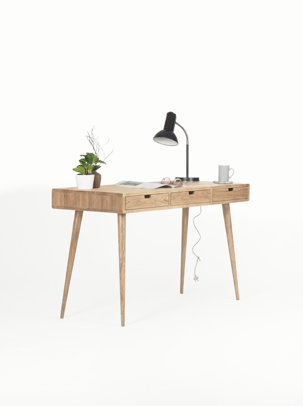 HEIM 140 writing desk with drawers in solid oak, angled view with a black lamp, an open magazine and a mug